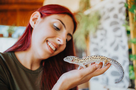 Smiling woman with red hair gently holding and observing a leopard gecko in her hand indoors, showcasing a love for reptiles and nature in a warm and inviting home environment.の写真素材