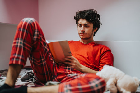 Young man reading a book in cozy plaid pajamas indoorsの写真素材