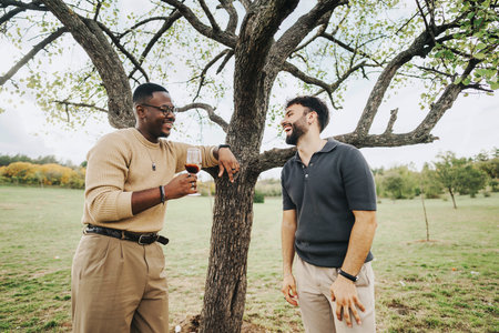 Two friends enjoying a laugh and wine under a large treeの写真素材
