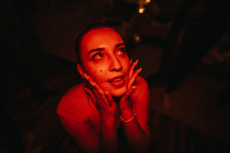 A young woman basked in red light, smiling with glitter on her face at a lively birthday...