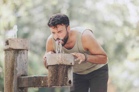 Man drinking water from a park fountain on a sunny day, capturing the essence of refreshment and outdoor activity.の写真素材