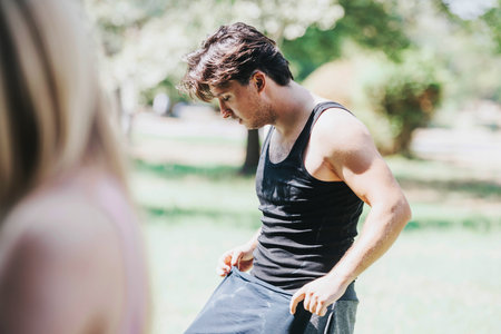 A young man takes a break after a workout in the park with friends. He is wearing a sleeveless shirt, enjoying the sunny day.の写真素材