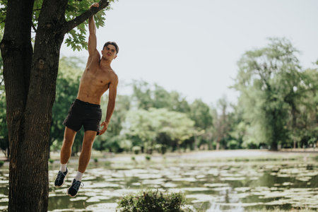 Shirtless man exercising by hanging from a tree branch in a scenic park settingの写真素材
