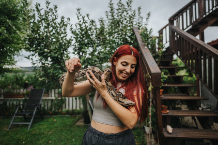 Red-haired girl joyfully holding a large snake outdoorsの写真素材