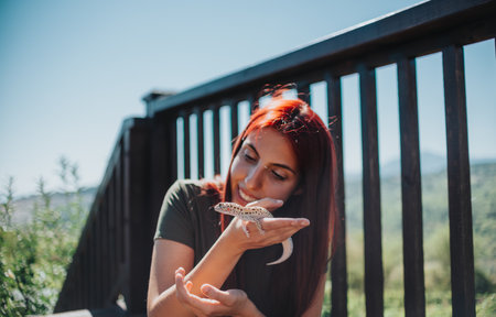 Young woman with red hair joyfully interacts with a small lizard outdoors on a sunny day, highlighting a connection with nature and enjoying a peaceful moment on a wooden deck.の写真素材