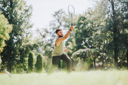 Young man enjoys playing badminton in a grassy park. The setting is sunny with lush green trees, capturing an active outdoor lifestyle.の写真素材