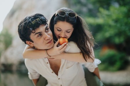 Joyful couple enjoying a playful moment outdoors, with the woman playfully biting an apple while on the man's back under the sunlight. They express happiness and carefree connection.の写真素材