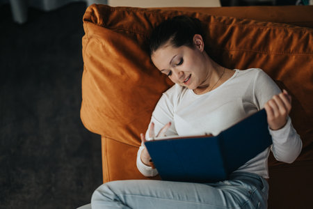Young woman relaxing on sofa while reading a book indoorsの写真素材