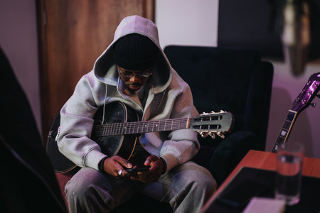 Young musician in hoodie texting while holding a guitar in cozy roomの写真素材