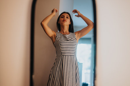 Confident woman in striped dress looks in the mirror at homeの写真素材
