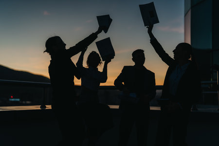 Silhouetted business people raising documents and celebrating success against a picturesque sunset.の写真素材