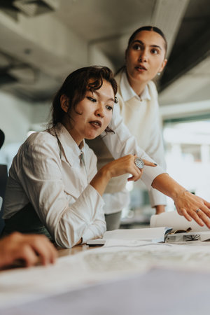 Two diverse businesswomen engage in a brainstorming session, analyzing documents and discussing...