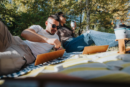 Young couple enjoying a relaxing day at the park with laptops and coffee on a sunny afternoon,...