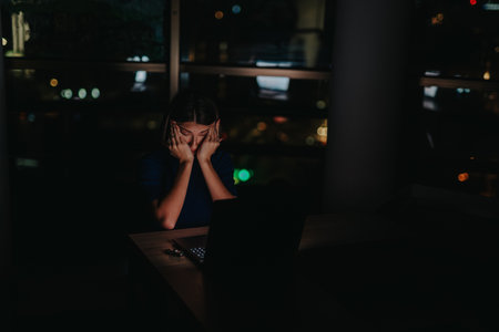 A business professional appears stressed while working late in a dimly lit office, illuminated by a laptop screen.の写真素材
