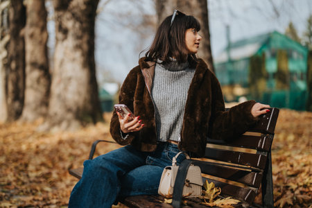Woman sitting on a park bench holding a phone during an autumn dayの写真素材
