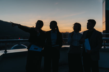 A group of business people stands on a rooftop at sunset, engaged in strategic discussion.の写真素材