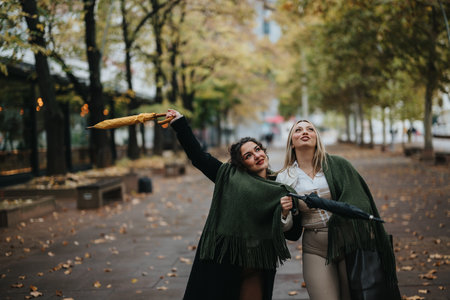 Two friends enjoying a stroll with umbrellas in an autumn urban parkの写真素材