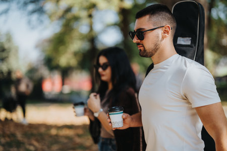 Young couple walking in park holding coffee cups and guitar case