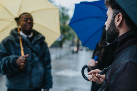 Diverse friends holding umbrellas during a rainy street discussion outdoorsの写真素材