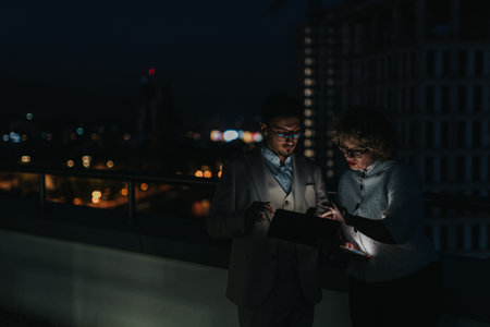 Two diverse people analyze project data on a tablet during a nighttime rooftop meeting.の写真素材