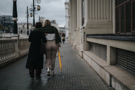 Two women walking closely on a city pathway with a yellow umbrella