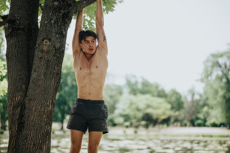 Shirtless man doing outdoor workout hanging from a tree branch in a parkの写真素材