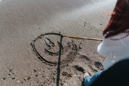 Person Drawing a Smiley Face on a Beach with a Stick in a Relaxed Settingの写真素材