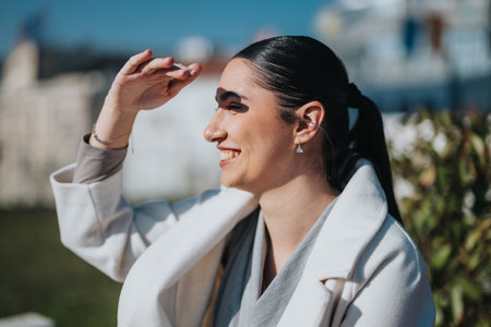 A confident woman with a casual outfit shielding her eyes from the sun while smiling outdoors.の写真素材