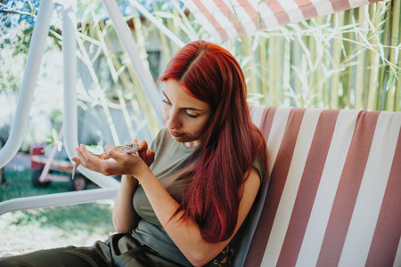 Girl with long red hair playing with a lizard on a striped garden swing on a sunny day