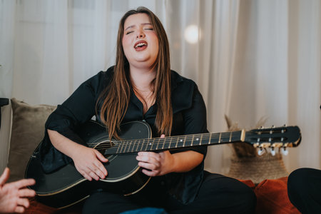 Woman joyfully singing and playing an acoustic guitar in a cozy settingの写真素材