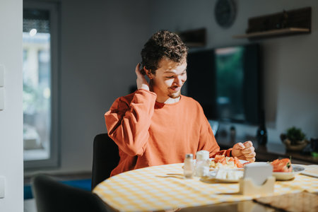 Young male teenager wearing an orange sweater sits at a table enjoying a meal with sliced fruits in a cozy modern interior.の写真素材