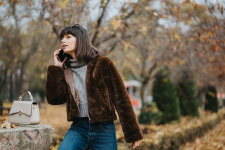 Woman in casual autumn attire talking on a phone in a park surrounded by fall foliageの写真素材