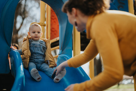 Child playing outdoors on a slide while a parent provides support and encouragement in a fun and active park settingの写真素材