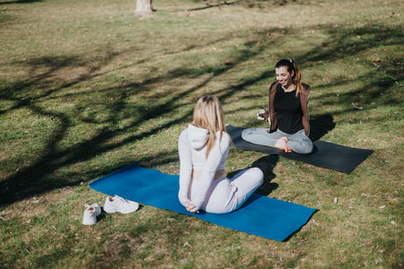 Two women practicing yoga outdoors while chatting in a sunny park settingの写真素材