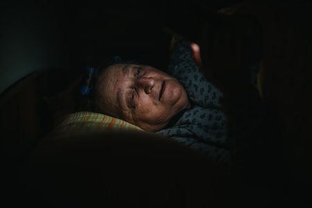 Senior woman lying in bed at night using a smart phone, illuminated by its screens lightの写真素材
