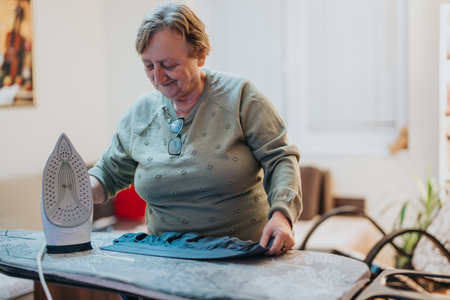 Elderly woman engaged in ironing clothes at home, showing focus and domestic careの写真素材
