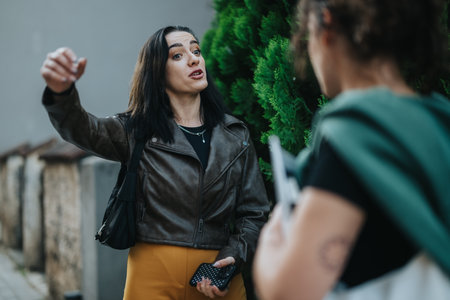 Two women discussing on a sidewalk next to greenery outdoors on a sunny dayの写真素材
