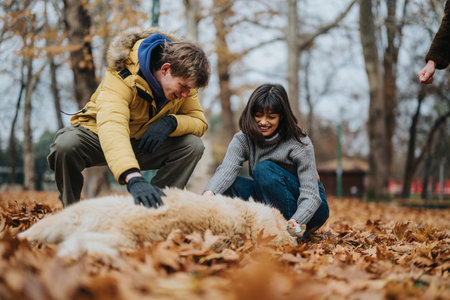 Young friends enjoying a playful moment outdoors in autumn while bonding with a fluffy dog amidst fallen leavesの写真素材