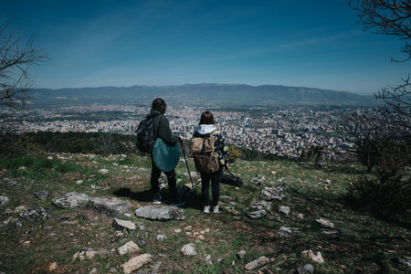 Two hikers exploring a scenic hilltop with a panoramic view of a sprawling city and distant mountain rangeの写真素材