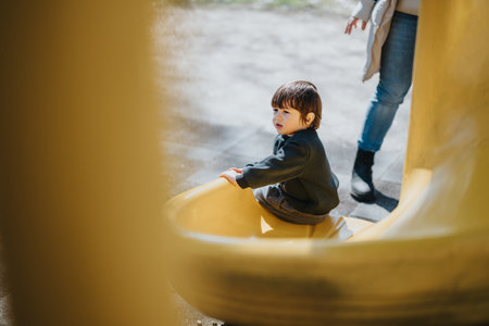 Young child sitting on a playground slide in a sunny park while interacting with an adultの写真素材