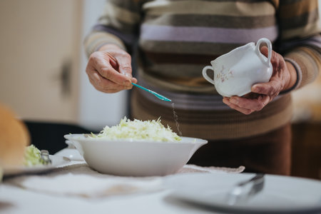 A senior woman preparing a meal by adding seasoning to a bowl of green salad.の写真素材