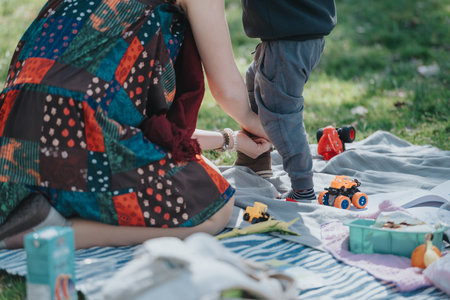 Parent and child bonding during an outdoor picnic activity, with toys and snacks spread on a blanket in a sunny park lot.の写真素材