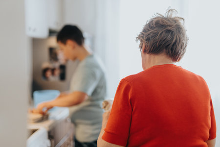 Elderly woman in red sweater with boy in kitchen setting a mealの写真素材