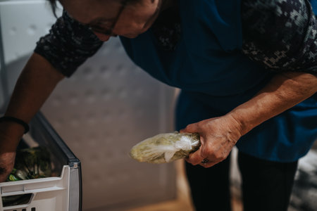 Elderly woman retrieving frozen vegetables from refrigerator compartment, highlighting daily home activityの写真素材