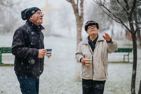 Two senior men enjoying snowfall at a park while holding beverage cups in winter clothingの写真素材