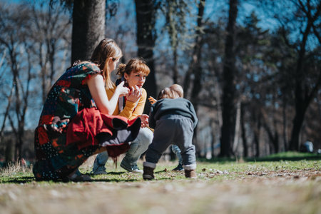 A candid park scene with two women interacting with a toddler under sunlit trees on a clear spring dayの写真素材