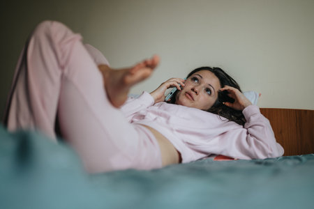 Young woman relaxing on bed while talking on phoneの写真素材