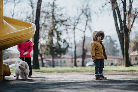 Child in a winter coat standing near a yellow slide with a dog beside them in a park on a chilly dayの写真素材