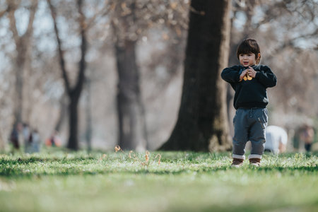 Young child standing on a green grassy park area during a sunny spring day surrounded by treesの写真素材