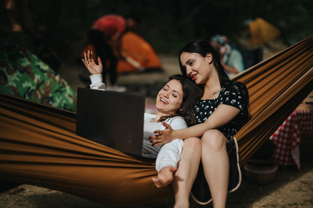 Two friends enjoying a laptop in a hammock during an outdoor camping tripの写真素材